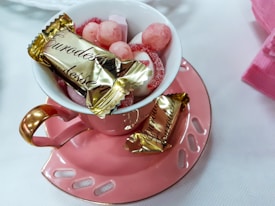 A pink teacup with a gold handle on a matching saucer holds a variety of sweets. The cup contains candies and chocolates, including a wrapped piece labeled 'Eurodessert'. The setting appears elegant with soft light, providing a cozy and inviting atmosphere.