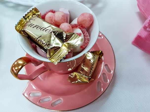 A pink teacup with a gold handle on a matching saucer holds a variety of sweets. The cup contains candies and chocolates, including a wrapped piece labeled 'Eurodessert'. The setting appears elegant with soft light, providing a cozy and inviting atmosphere.