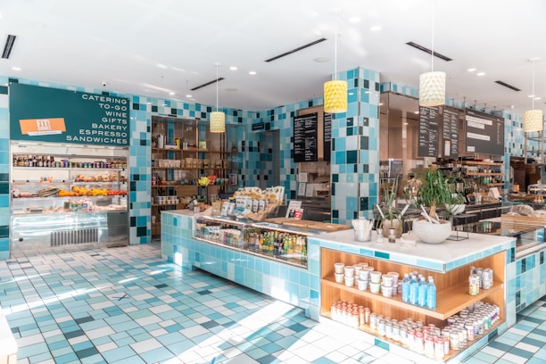 A modern and bright cafe interior featuring turquoise and white tiled walls and floors. The space includes shelves stocked with various goods, a counter displaying snacks and drinks, and menu boards hanging above. Plants in stylish pots add a touch of greenery to the decor. Hanging pendant lights illuminate the area, while a sign lists catering, wine, gifts, bakery, espresso, and sandwiches.