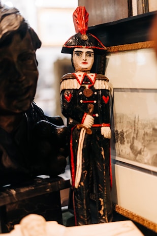 A carved wooden figurine of a soldier is prominently displayed with black and red detailing. The figure wears a period uniform with decorative elements such as epaulettes and a tall hat with a red plume. It stands beside a framed black-and-white sketch or photograph. A darker bust sculpture and another indistinct object are partially visible beside the main figurine.