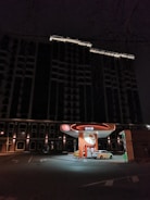 A gas station with a bright orange and white color scheme is illuminated in an urban setting during nighttime. A white car is parked at one of the fueling stations beneath a dark, tall building. The area is mostly quiet with a few streetlights around.