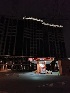 A gas station with a bright orange and white color scheme is illuminated in an urban setting during nighttime. A white car is parked at one of the fueling stations beneath a dark, tall building. The area is mostly quiet with a few streetlights around.
