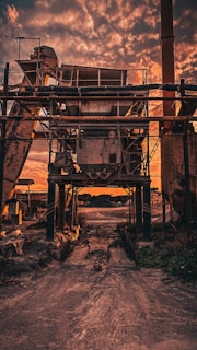 An industrial structure looms in the foreground with a complex framework of metal beams and machinery. The scene is set against a dramatic sky filled with vivid orange and purple clouds. The ground appears worn and dusty, evoking a sense of dereliction and abandonment.