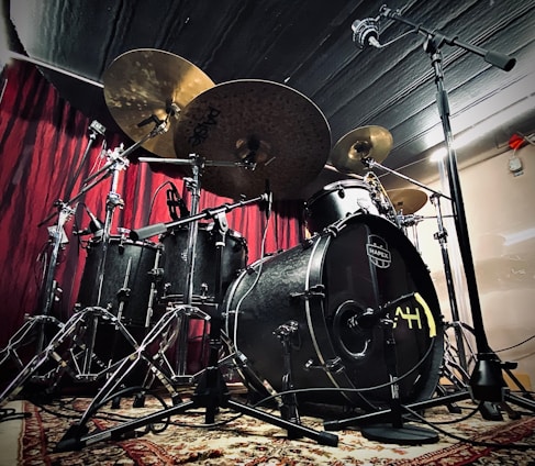 A drum set is positioned against a red curtain backdrop, featuring multiple cymbals, a bass drum, tom-toms, and a hi-hat. The drum kit is adorned with metallic hardware and set on a patterned rug. Overhead and floor-mounted microphones are set up around the drum kit, suggesting a recording or performance setting.