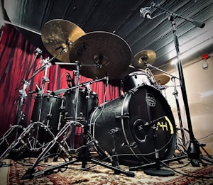 A drum set is positioned against a red curtain backdrop, featuring multiple cymbals, a bass drum, tom-toms, and a hi-hat. The drum kit is adorned with metallic hardware and set on a patterned rug. Overhead and floor-mounted microphones are set up around the drum kit, suggesting a recording or performance setting.
