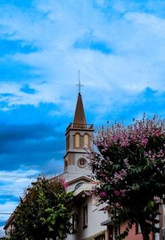 A church steeple with a cross on top stands prominently against a vibrant blue sky. Surrounding the building are lush trees adorned with purple flowers, adding a touch of color and life to the scene.