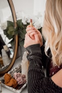 A person with blonde hair is holding a lipstick in front of a decorative mirror. The setting includes a tray with crystals and a bottle, while white flowers are visible in the reflection.