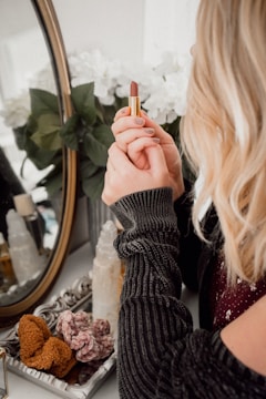 A person with blonde hair is holding a lipstick in front of a decorative mirror. The setting includes a tray with crystals and a bottle, while white flowers are visible in the reflection.