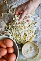 A hand holding freshly made pasta on a floured wooden surface, surrounded by a bowl of brown eggs and a bowl of flour.