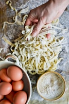 A hand holding freshly made pasta on a floured wooden surface, surrounded by a bowl of brown eggs and a bowl of flour.