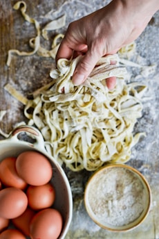 A hand holding freshly made pasta on a floured wooden surface, surrounded by a bowl of brown eggs and a bowl of flour.