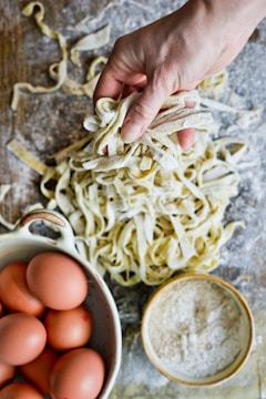 A hand holding freshly made pasta on a floured wooden surface, surrounded by a bowl of brown eggs and a bowl of flour.