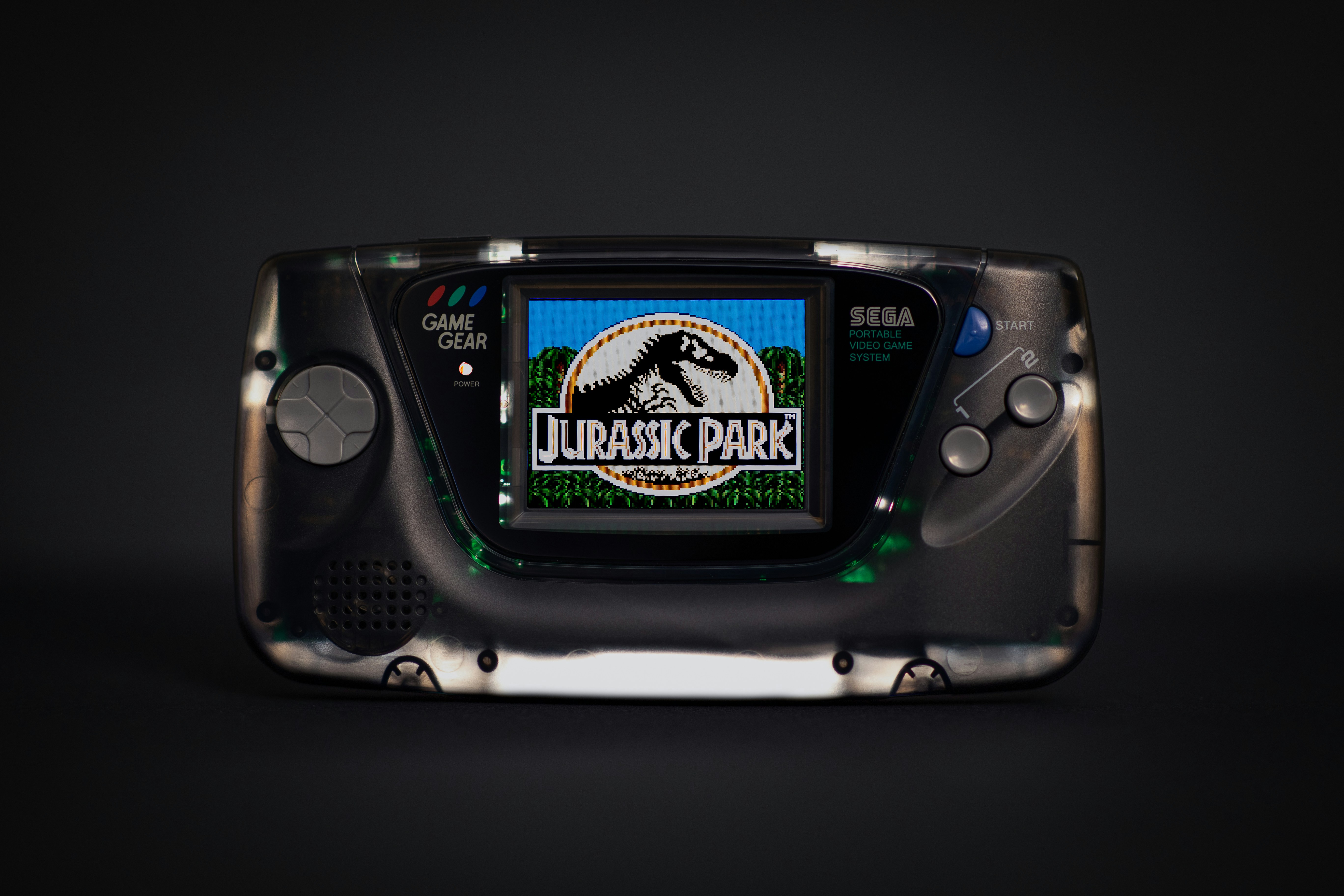 A retro backlit Sega Game Gear with Jurassic Park game running, black background. Semi transparent black shell and McWill LCD display screen upgrade.