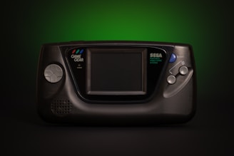 A vintage Sega Game Gear handheld video game console, prominently featuring a large screen in the center. The console has a directional pad on the left and two start buttons on the right. The unit is primarily black with subtle branding in white text. The background fades from dark to a vibrant green which adds a retro aesthetic.