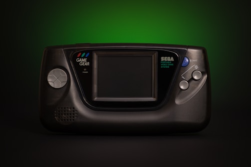 A vintage Sega Game Gear handheld video game console, prominently featuring a large screen in the center. The console has a directional pad on the left and two start buttons on the right. The unit is primarily black with subtle branding in white text. The background fades from dark to a vibrant green which adds a retro aesthetic.