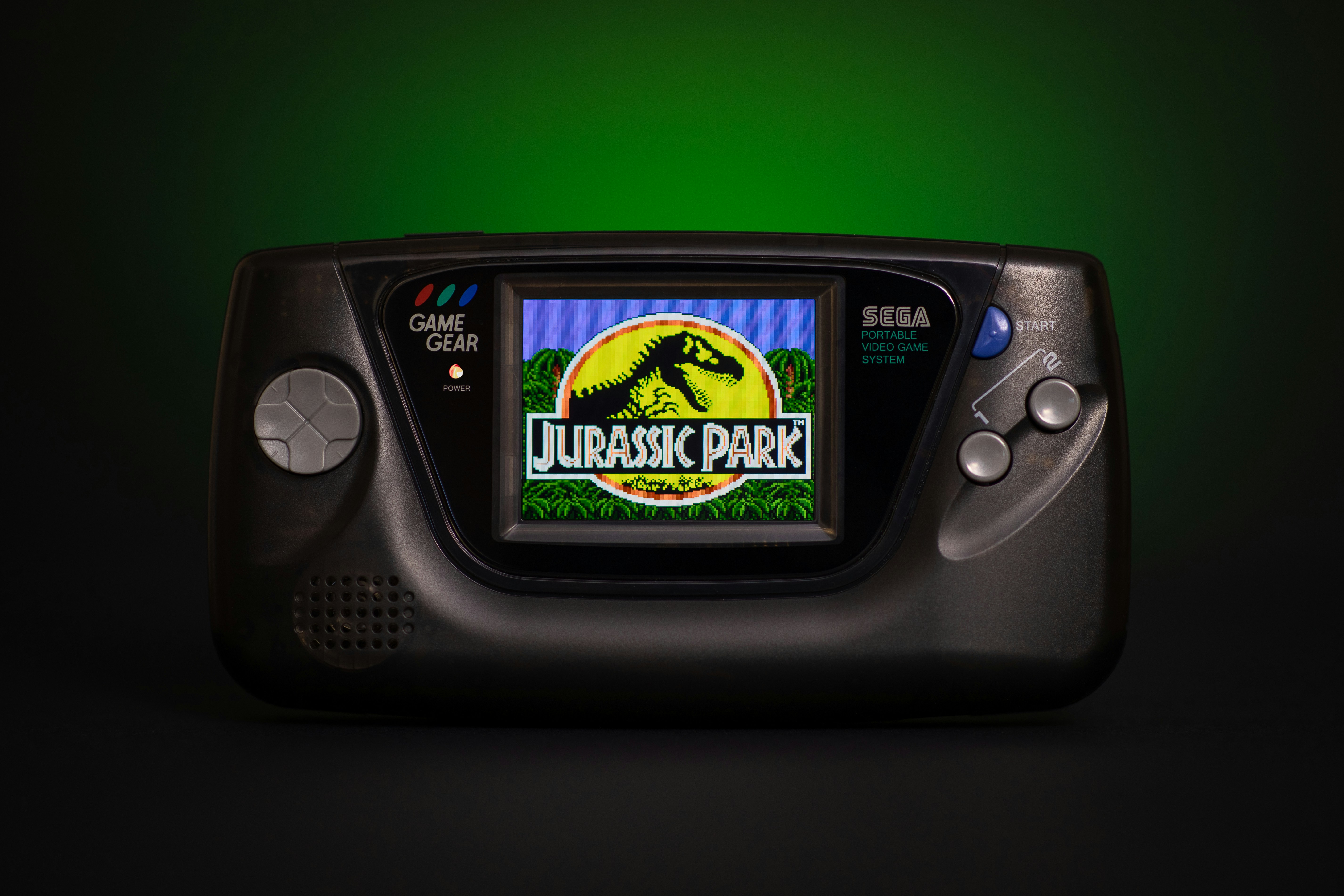 A retro Sega Game Gear with Jurassic Park game running, black background with a green glow. Semi transparent black shell and McWill LCD display screen upgrade.