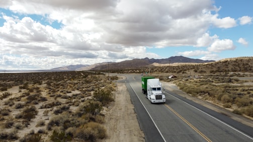 A white semi-truck with a green trailer is traveling on a lonely highway that winds through a barren desert landscape. The sky above is filled with white clouds, casting shadows over the sandy terrain and sparse vegetation. In the distance, low mountains are visible beneath the cloudy sky.