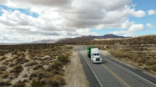 A white semi-truck with a green trailer is traveling on a lonely highway that winds through a barren desert landscape. The sky above is filled with white clouds, casting shadows over the sandy terrain and sparse vegetation. In the distance, low mountains are visible beneath the cloudy sky.