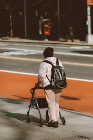 A person wearing a pink outfit and black hat is walking on a sidewalk using a walker. They have a black backpack and appear to be moving towards a crosswalk on a street. There is a red and grey telephone booth or similar structure in the background.