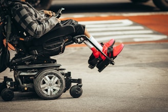 A person is seated in a motorized wheelchair on a city street, wearing bright red sneakers and a patterned hoodie. The wheels of the wheelchair and the urban environment are visible.