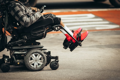 A person is seated in a motorized wheelchair on a city street, wearing bright red sneakers and a patterned hoodie. The wheels of the wheelchair and the urban environment are visible.