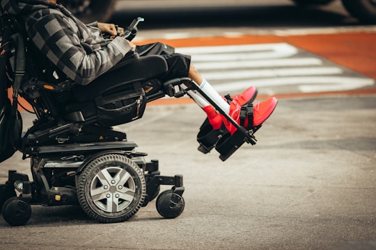 A person is seated in a motorized wheelchair on a city street, wearing bright red sneakers and a patterned hoodie. The wheels of the wheelchair and the urban environment are visible.