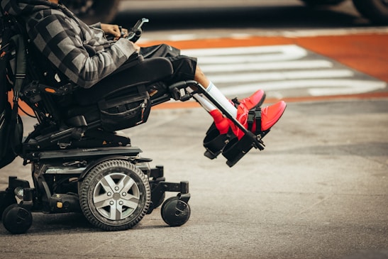 A person is seated in a motorized wheelchair on a city street, wearing bright red sneakers and a patterned hoodie. The wheels of the wheelchair and the urban environment are visible.