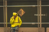 A person wearing a bright yellow reflective jacket with a hood stands near a construction site. They are leaning against a concrete barrier and are speaking on a mobile phone. The background features a green wooden board with a 'POST NO BILLS' sign and metal scaffolding.