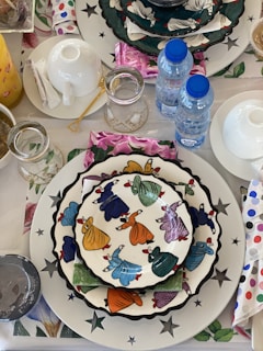 A colorful table setting features stacked plates with whimsical patterns of dancing figures in vibrant clothing, surrounded by various tableware including cups, glasses, and water bottles. The table is adorned with a patterned tablecloth with floral and star motifs, along with napkins with polka dots.