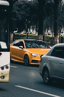 A bright yellow car is driving on a tree-lined road, surrounded by other vehicles in traffic. The scene includes a white bus on the left and a gray car on the right. There are tall trees and faintly visible foliage in the background.