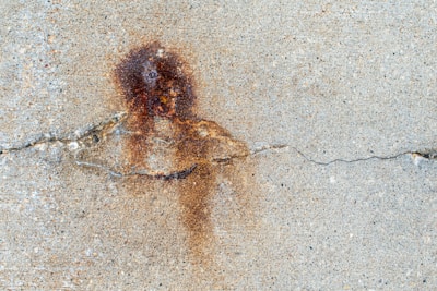 A textured concrete surface with a prominent brownish rust stain near the center. The rust mark is irregular in shape and coloration, surrounded by lighter concrete with a visible crack running horizontally across the image.