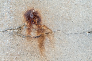 A textured concrete surface with a prominent brownish rust stain near the center. The rust mark is irregular in shape and coloration, surrounded by lighter concrete with a visible crack running horizontally across the image.