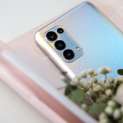 A smartphone with a shimmering back panel and multiple camera lenses is placed on a surface, partially adorned with small white flowers. The image features a soft, light setting with blurred floral accents in the foreground.