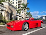 A shiny red convertible parked along the sunny California coast with palm trees in the background.