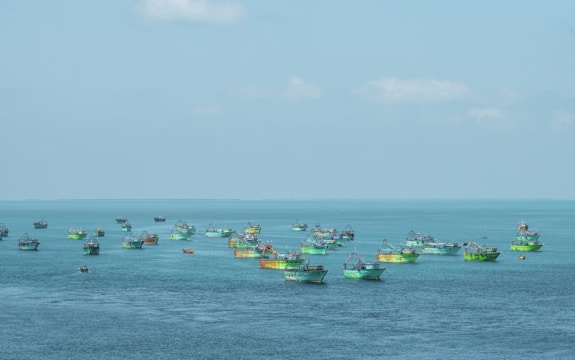 Numerous fishing boats with vibrant green and blue colors are spread across a vast expanse of calm ocean waters, under a clear blue sky.