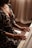 A person is playing the piano, with hands gracefully positioned on the keys. The individual is wearing a patterned dress and is seated in a softly lit room with sheer curtains in the background.