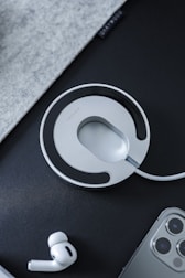 A circular charging pad with a sleek design and a modern aesthetic is featured prominently. It is placed on a dark surface alongside a pair of wireless earbuds and the corner of a smartphone with triple cameras. The setting is minimalistic with an emphasis on technology and modern gadgets.