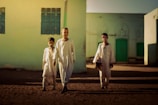 Three young boys walk together across a sunlit courtyard with light sand-colored walls and green doors. They wear loose-fitting traditional clothing and cast long shadows on the ground.