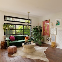 A well-decorated living room with a large green velvet sofa adorned with colorful cushions, positioned in front of a big window with black frames. The room features an eclectic mix of artwork on the walls, a contemporary chandelier, and various indoor plants adding a touch of greenery. A round wooden coffee table sits atop a beige rug, surrounded by other decor items, including a unique wooden side table and a leather pouf. Natural light floods the room, creating a bright and welcoming atmosphere.