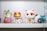 A collection of colorful figurines, each with distinct features and designs, is arranged on a white shelf. The central figure resembles a maneki-neko, featuring a shiny gold face with a white cat costume. On the left, there is a whimsical character with elfin ears holding an object resembling a pink seashell with purple accents. Nearby, another figure resembles a playful cat-like creature with large eyes and contrasting orange and white fur patterns. In the background, partially obscured characters add to the eclectic mix of toys.