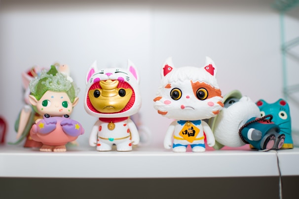 A collection of colorful figurines, each with distinct features and designs, is arranged on a white shelf. The central figure resembles a maneki-neko, featuring a shiny gold face with a white cat costume. On the left, there is a whimsical character with elfin ears holding an object resembling a pink seashell with purple accents. Nearby, another figure resembles a playful cat-like creature with large eyes and contrasting orange and white fur patterns. In the background, partially obscured characters add to the eclectic mix of toys.
