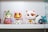A collection of colorful figurines, each with distinct features and designs, is arranged on a white shelf. The central figure resembles a maneki-neko, featuring a shiny gold face with a white cat costume. On the left, there is a whimsical character with elfin ears holding an object resembling a pink seashell with purple accents. Nearby, another figure resembles a playful cat-like creature with large eyes and contrasting orange and white fur patterns. In the background, partially obscured characters add to the eclectic mix of toys.