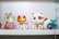 A collection of colorful figurines, each with distinct features and designs, is arranged on a white shelf. The central figure resembles a maneki-neko, featuring a shiny gold face with a white cat costume. On the left, there is a whimsical character with elfin ears holding an object resembling a pink seashell with purple accents. Nearby, another figure resembles a playful cat-like creature with large eyes and contrasting orange and white fur patterns. In the background, partially obscured characters add to the eclectic mix of toys.