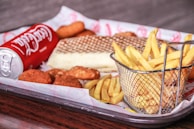 A tray holds a selection of fast food items including a can of Coca-Cola, a basket of french fries, a sandwich with a waffle-like texture on the bread, and several breaded chicken nuggets.