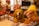 A golden retriever sits attentively with a happy expression in a living room setting. The dog is surrounded by traditional-style wooden chairs with red cushions. In the foreground, a blurred hand is holding a piece of food, possibly intended for the dog.