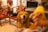 A golden retriever sits attentively with a happy expression in a living room setting. The dog is surrounded by traditional-style wooden chairs with red cushions. In the foreground, a blurred hand is holding a piece of food, possibly intended for the dog.