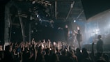 A live concert scene featuring a band performing on a stage. The lead singer is holding a microphone, engaging with the audience, while the guitarist and drummer are also visible. A large crowd is in the foreground, with many people raising their hands and holding up phones to capture the moment. The stage is illuminated with various lighting, creating a dynamic atmosphere.