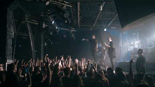 A live concert scene featuring a band performing on a stage. The lead singer is holding a microphone, engaging with the audience, while the guitarist and drummer are also visible. A large crowd is in the foreground, with many people raising their hands and holding up phones to capture the moment. The stage is illuminated with various lighting, creating a dynamic atmosphere.