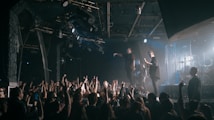 A live concert scene featuring a band performing on a stage. The lead singer is holding a microphone, engaging with the audience, while the guitarist and drummer are also visible. A large crowd is in the foreground, with many people raising their hands and holding up phones to capture the moment. The stage is illuminated with various lighting, creating a dynamic atmosphere.