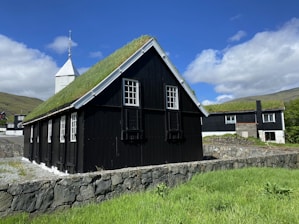 A traditional building with a grass-covered roof stands against a backdrop of vibrant green grass and a bright blue sky dotted with clouds. The building has black wooden walls and white framed windows, creating a stark contrast with the lush surrounding landscape. Another structure is visible in the background.